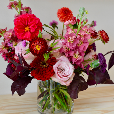 A vibrant flower arrangement bursting with color! This bouquet features red zinnias, pink stock, and dahlias in shades of red and orange, accented with purple and burgundy foliage.  Perfect for birthdays, anniversaries, or any celebration. Available for local delivery in Wake Forest, NC.